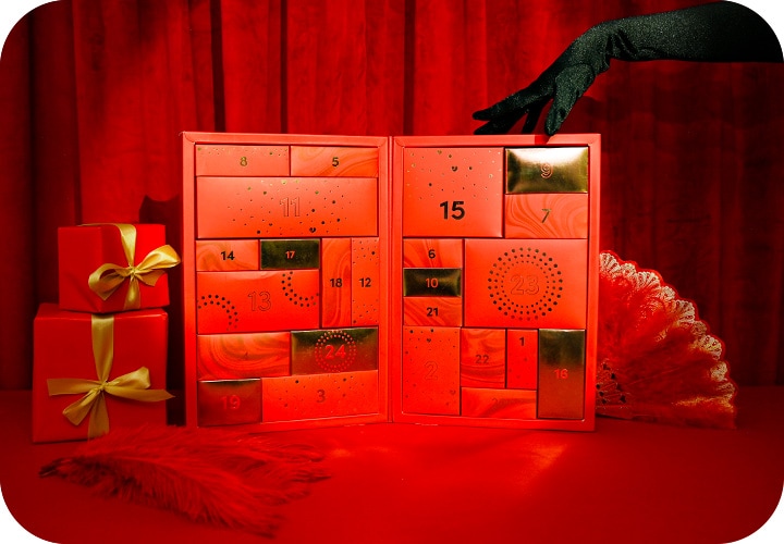 [La boite du calendrier de l'avent espaceplaisir 2024, placée dans un décor entièrement rouge. Une main habillée d'un long gant noir et pailleté est poséee sur la boîte.]